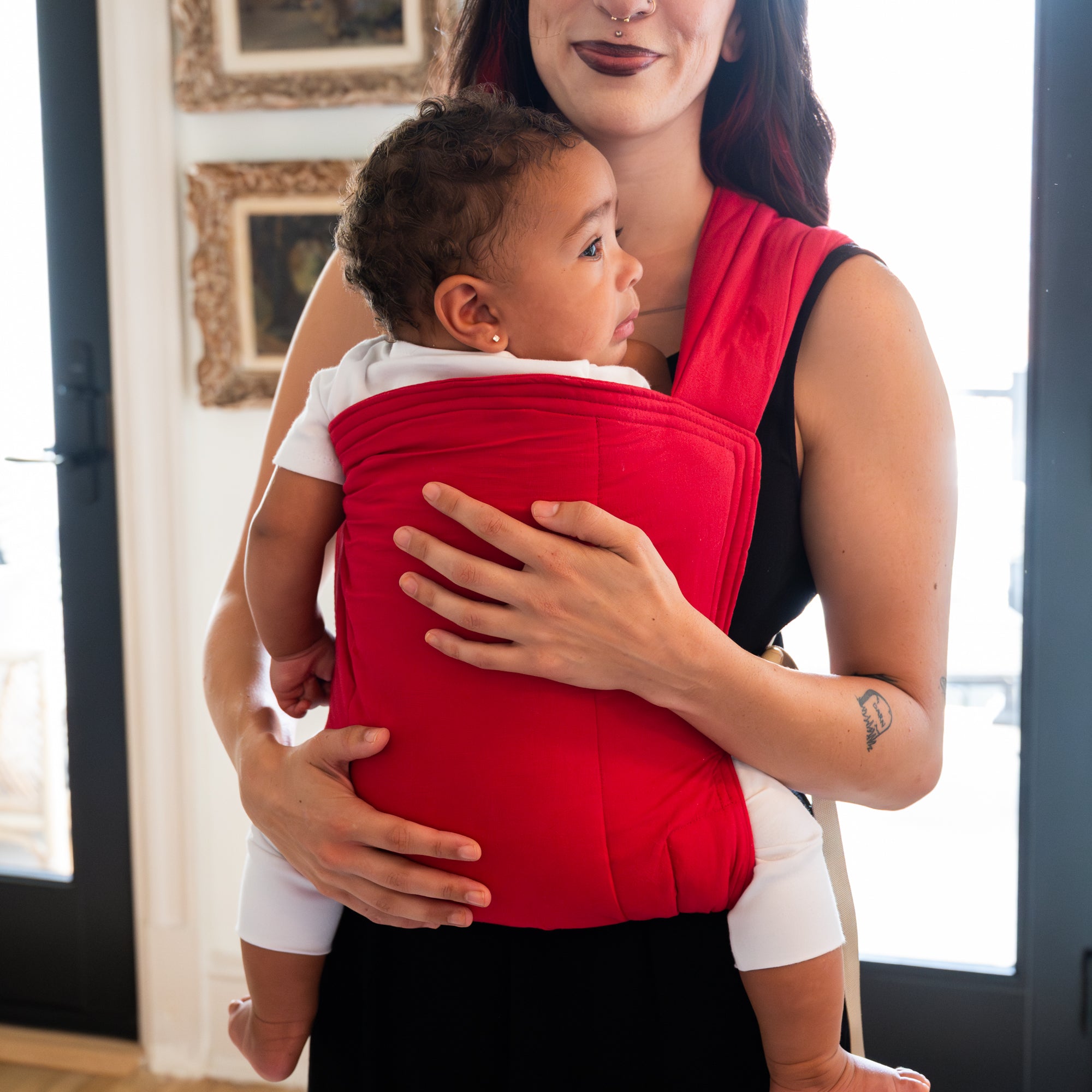Woman holding a baby in a red soft carrier indoors.
