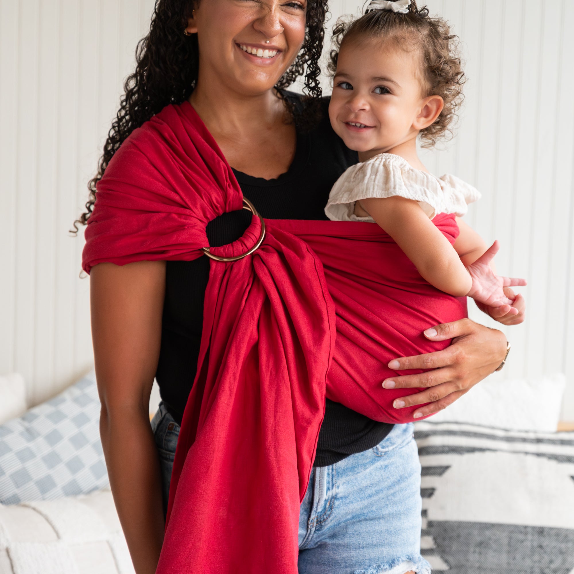 Woman holding a child in a red sling baby carrier against a neutral background