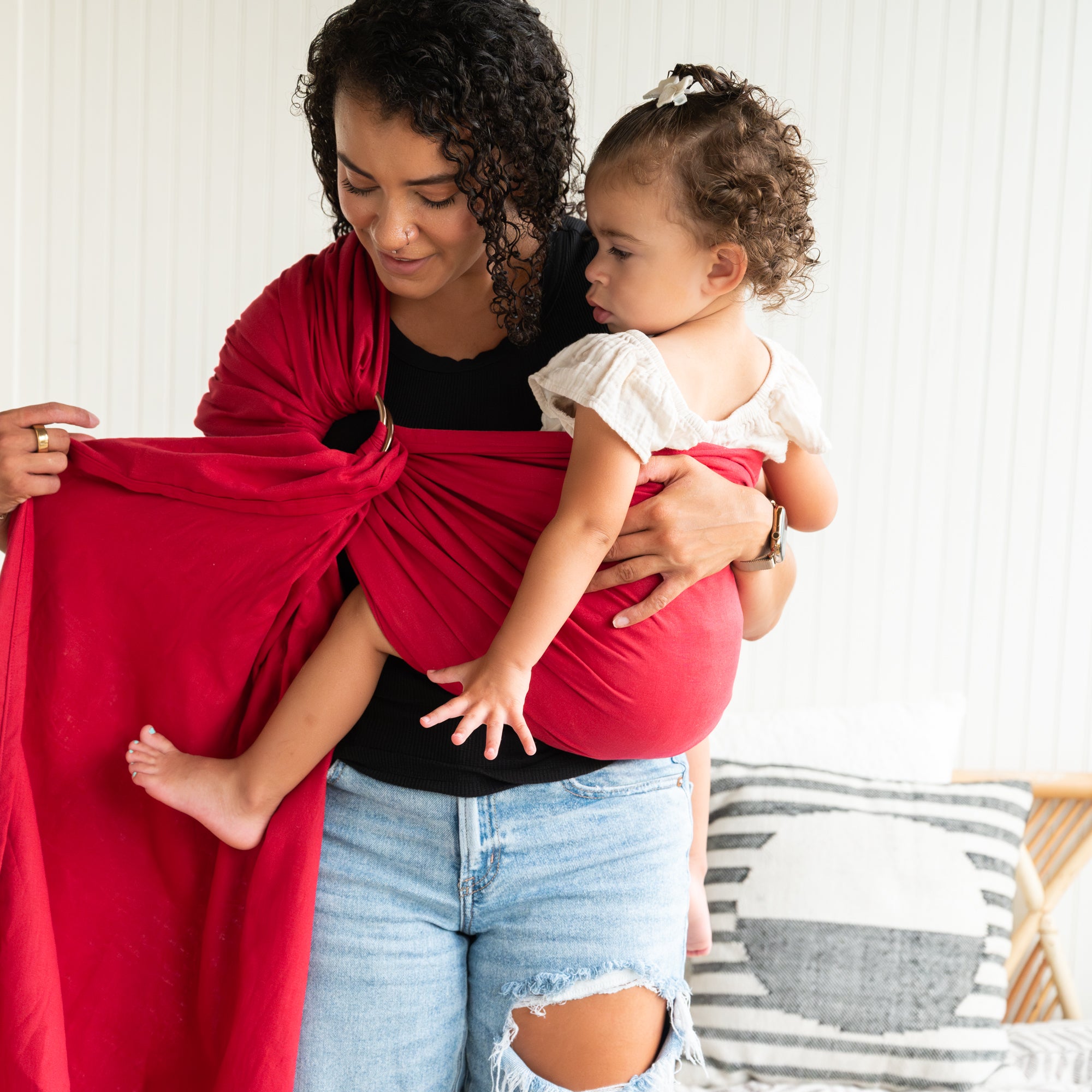Woman holding a child in a red baby wrap against a white wall.