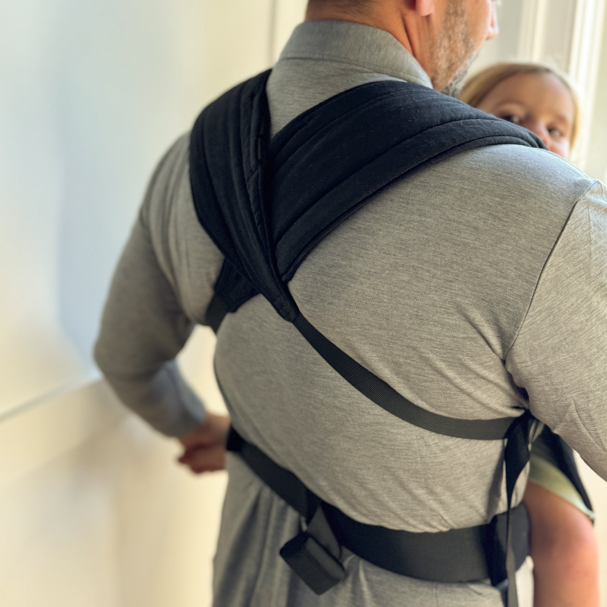 Man carrying a child in a black baby carrier against a light-colored wall.