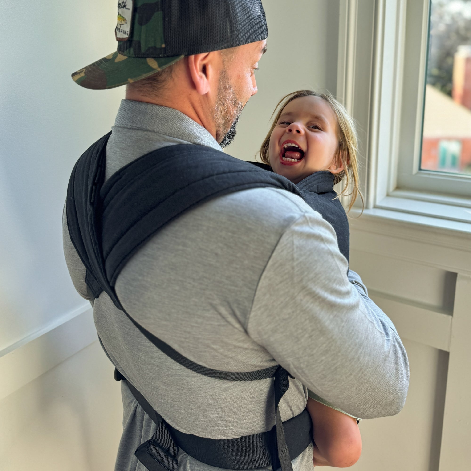 Man carrying a child in a black baby carrier against a light-colored wall.