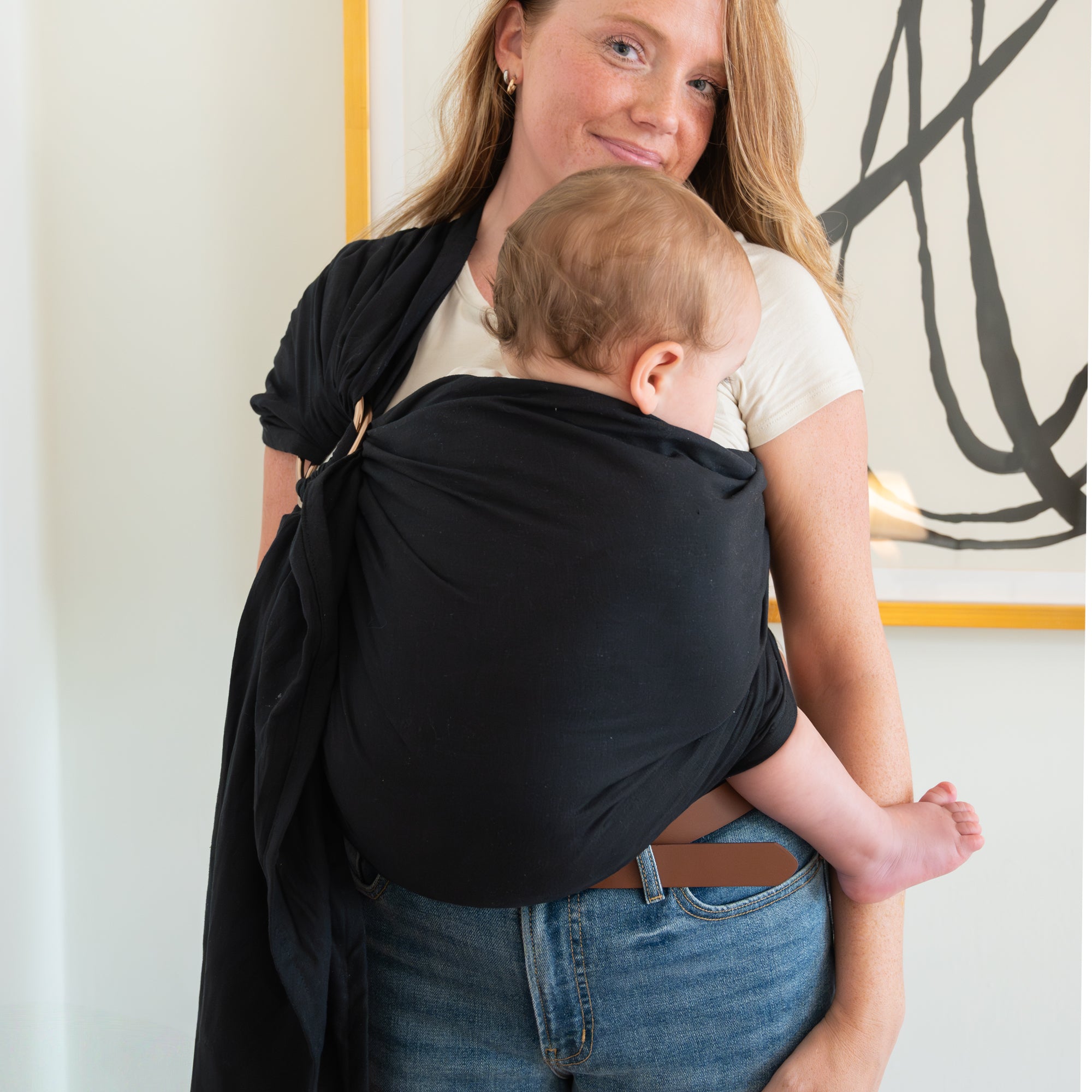 Woman carrying a baby in a black sling against a light-colored wall.