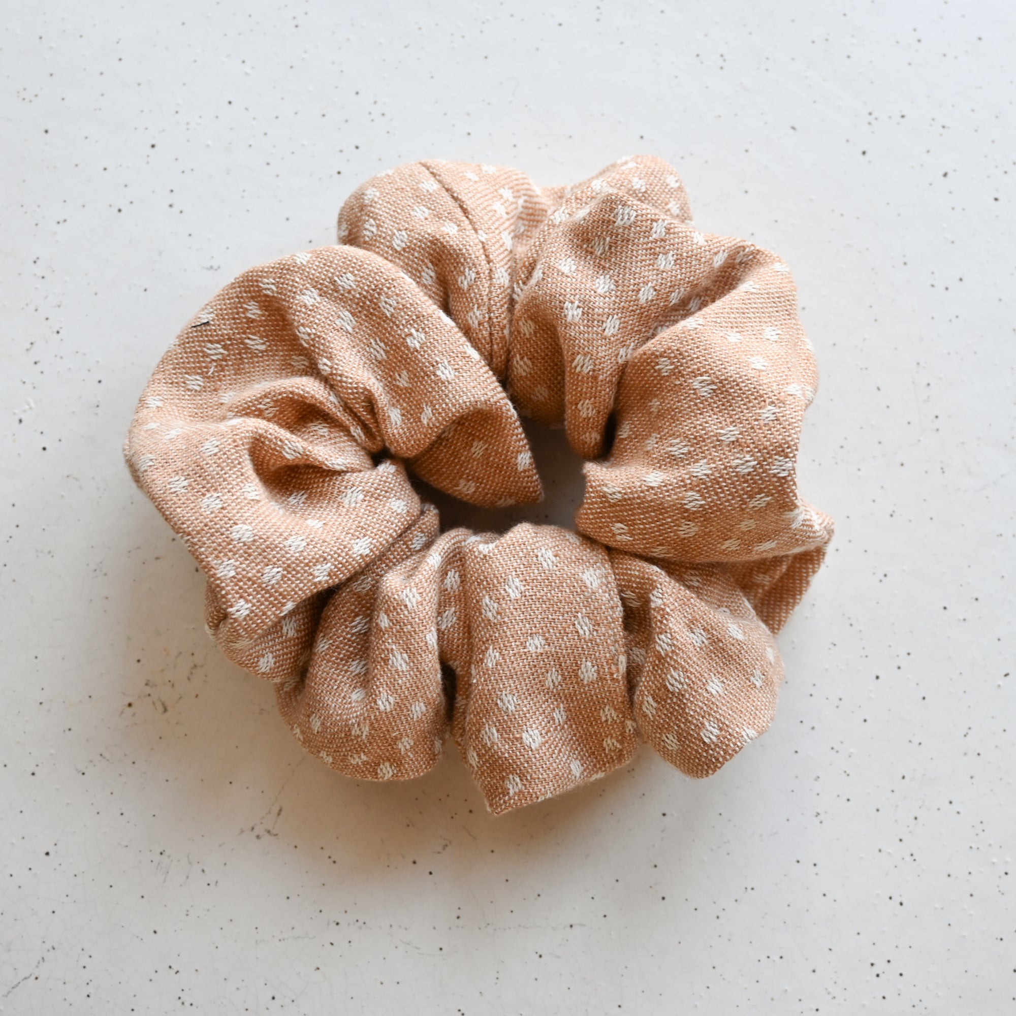 A single oversized scrunchie in warm camel woven fabric with a small scattered white dash or spot pattern woven into the textile.