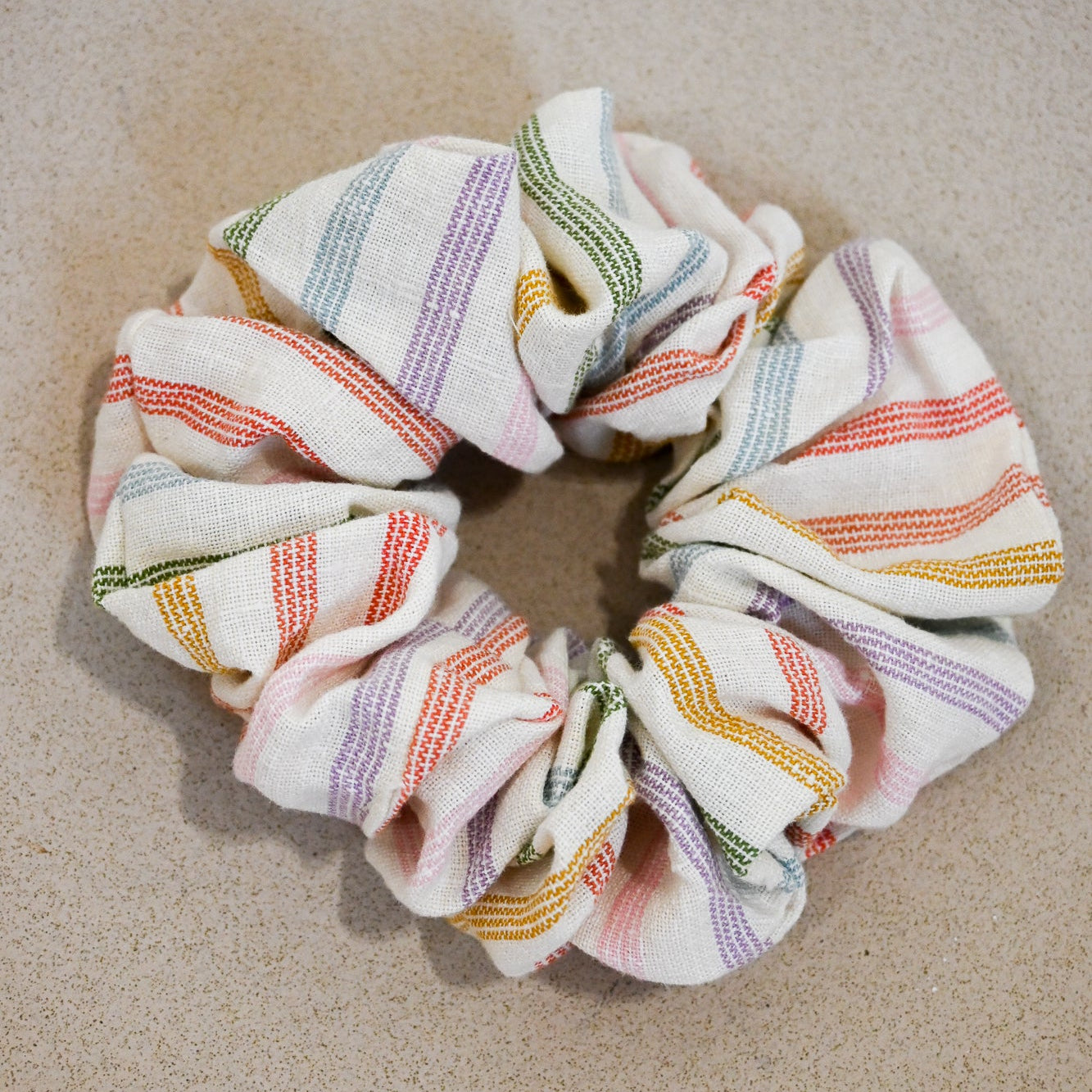 A single oversized scrunchie in natural cream woven fabric with narrow multicolour rainbow stripes in coral, mustard, green, lilac, pink, and pale blue.