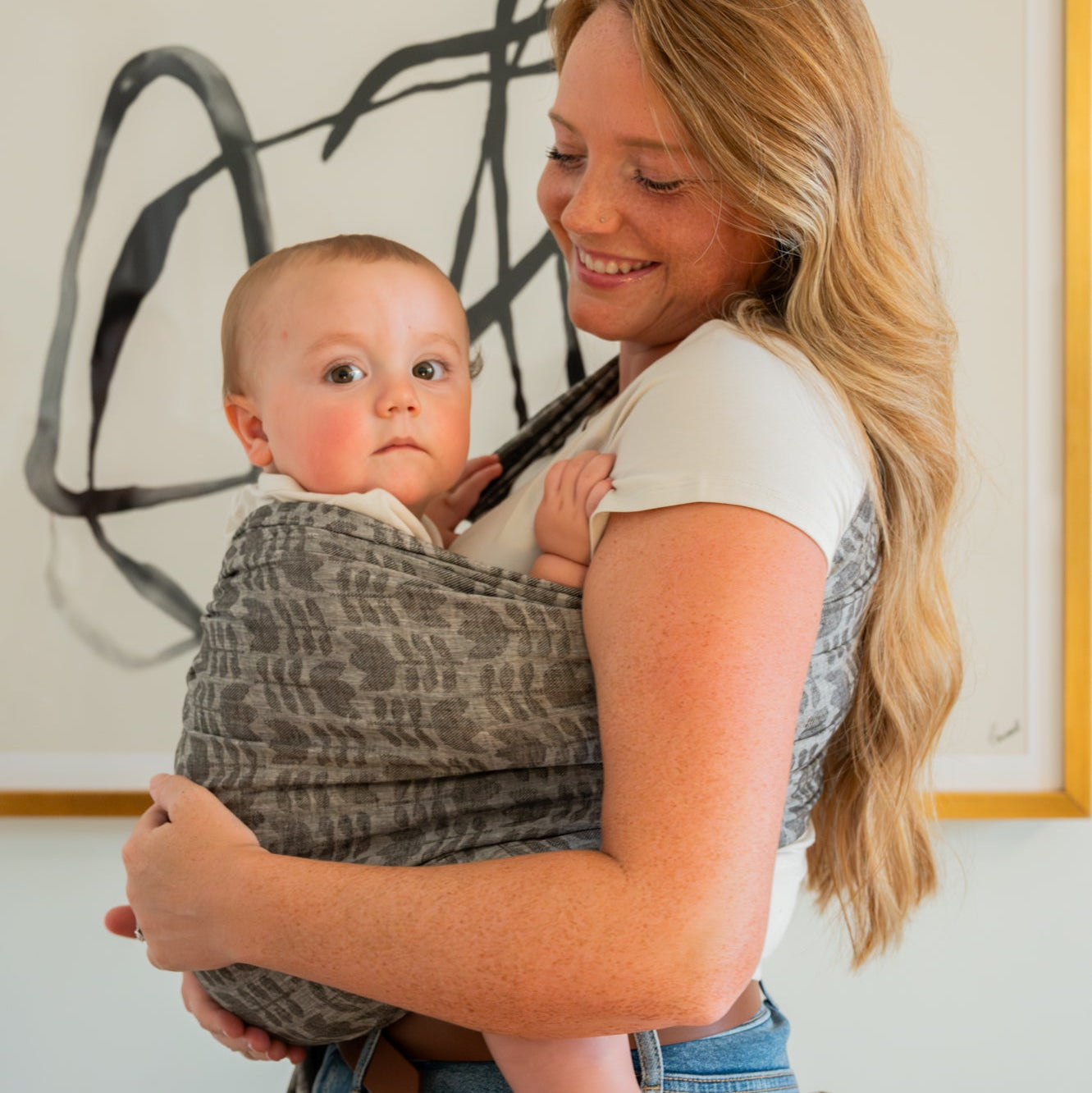 Woman holding a baby in a sling against a white wall with abstract art.