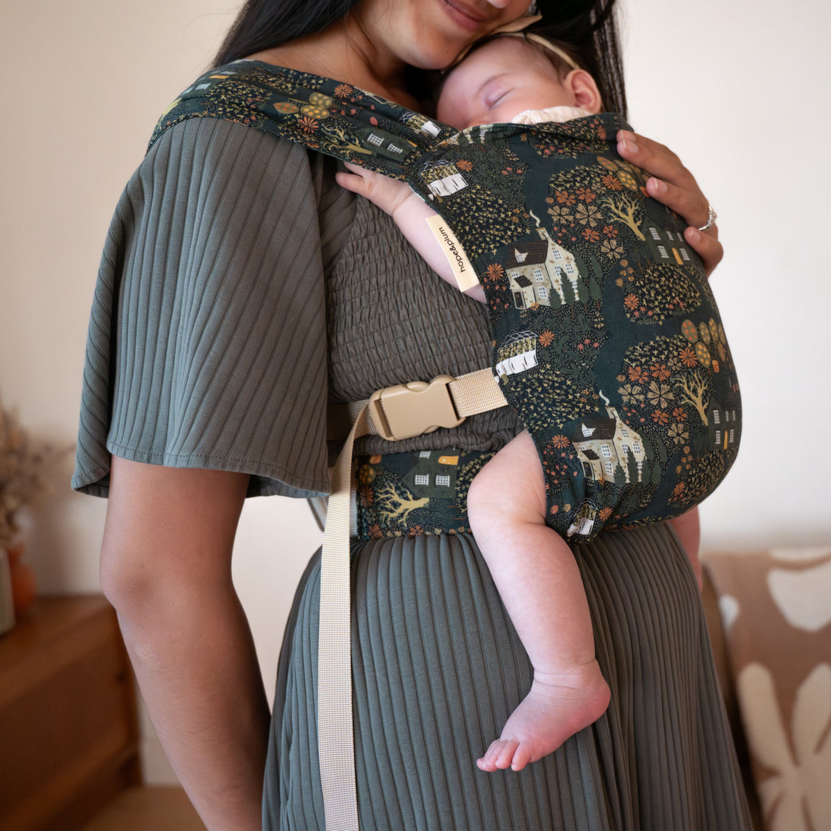 A caregiver nuzzles a sleeping baby in the Sprout in Village — a deep green woven fabric printed with a whimsical folk-art scene of cottages, glowing trees, and autumn florals.