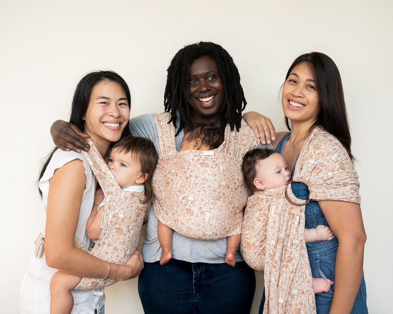 Three smiling caregivers each carry a baby in matching Woodland print carriers — a warm blush woven fabric with detailed botanical and mushroom illustrations — wearing a Lark, a baby wrap, and a ring sling.