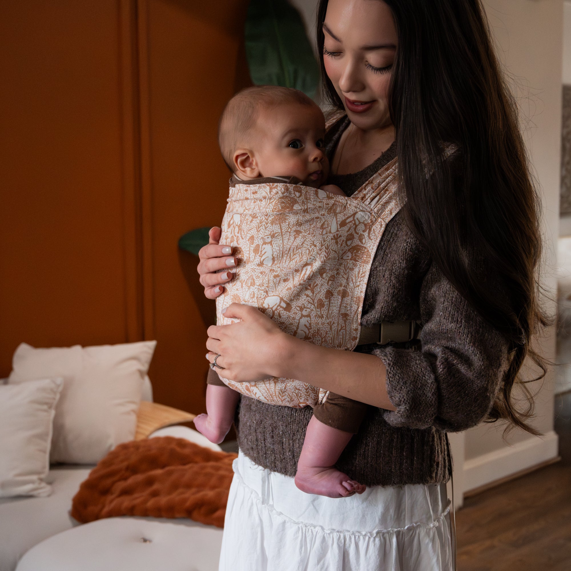 Woman holding a baby in a white carrier with brown forest animals in a cozy indoor setting