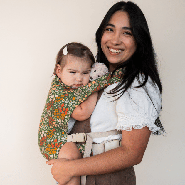 A smiling caregiver holds a grumpy-faced baby clutching a stuffed bear in the Fête carrier — a rich green woven fabric densely printed with orange, yellow, and white wildflowers and strawberries.