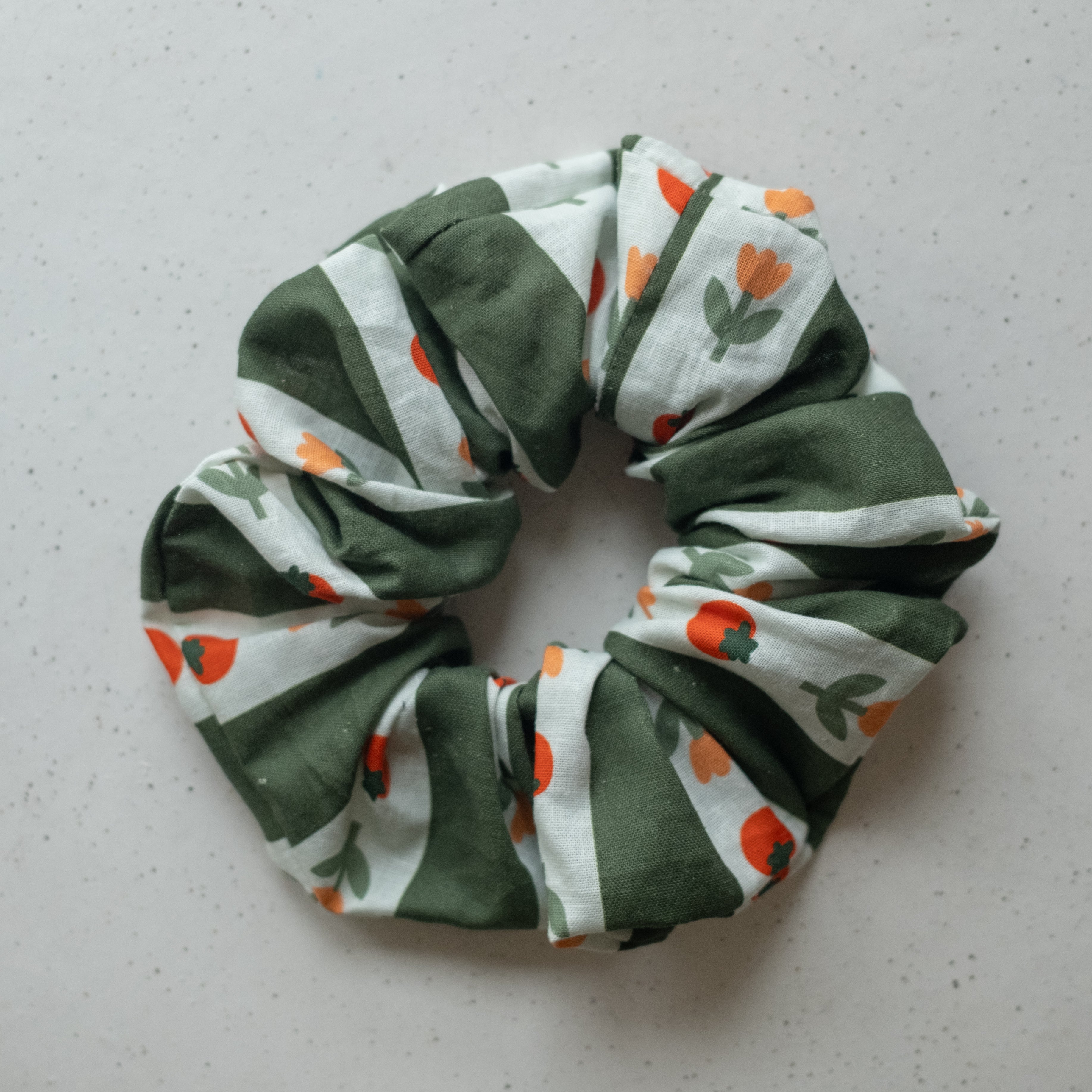 A single oversized scrunchie in olive green and white wide-stripe woven fabric printed with small orange and red tomatoes and green stems and leaves.