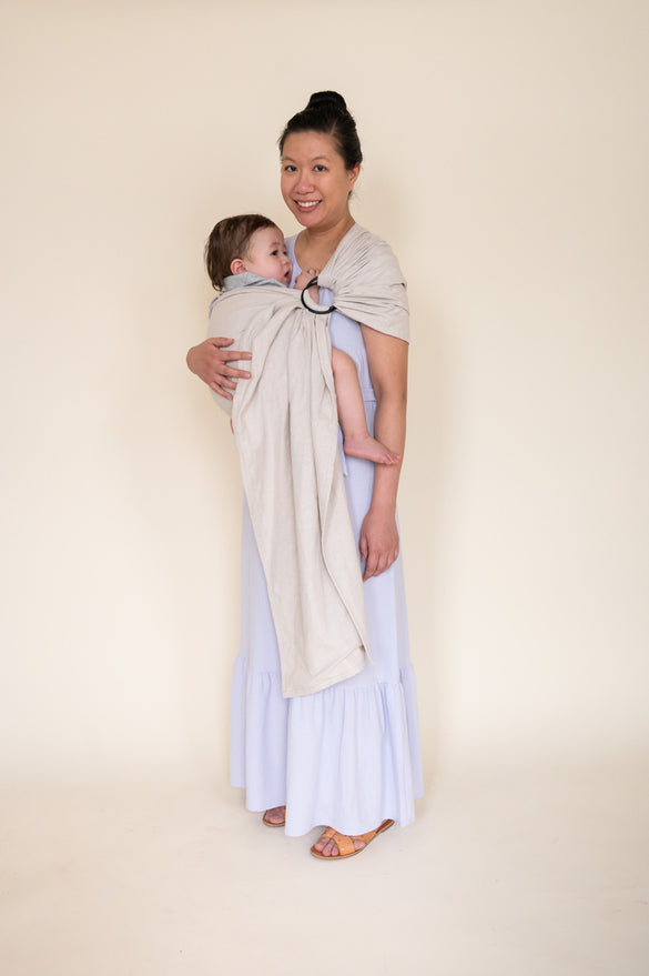 Asian mother holding her child in a light gray ring sling baby carrier. 