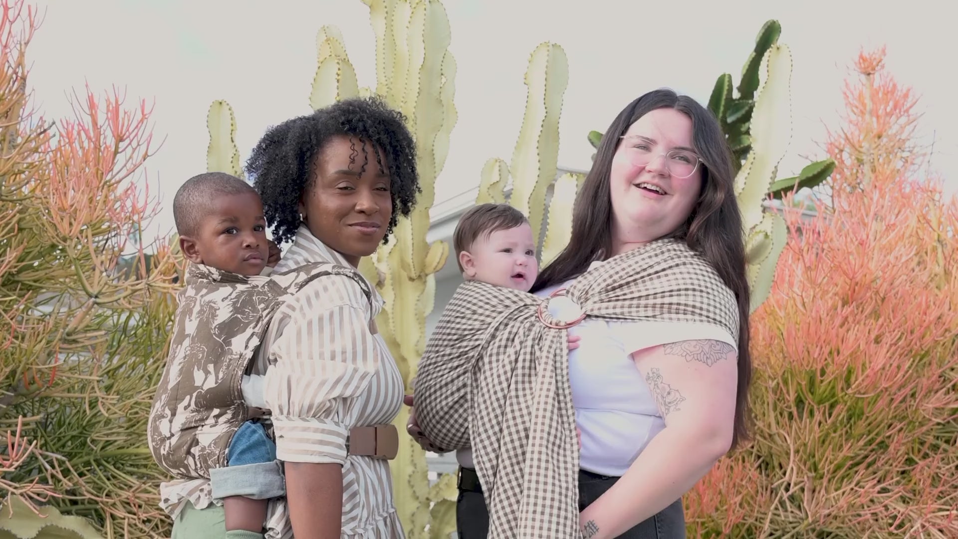 Editorial video for the Rodeo and Clove baby carrier collection, showing both carriers worn outdoors and indoors: two women carrying a toddler and baby in the horse-print Rodeo carrier and gingham Clove ring sling by a cactus garden, plus close-ups of a woman in a cowboy hat and a father wearing the carriers with their children.