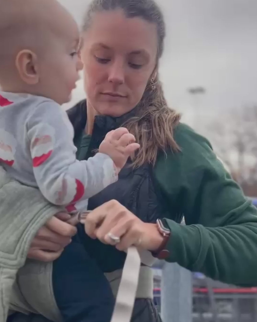 A woman stands in a parking lot wearing a baby in a Hope & Plum Lark buckle carrier in Palm green, fastening the waist strap and keeping the baby snug against her chest.