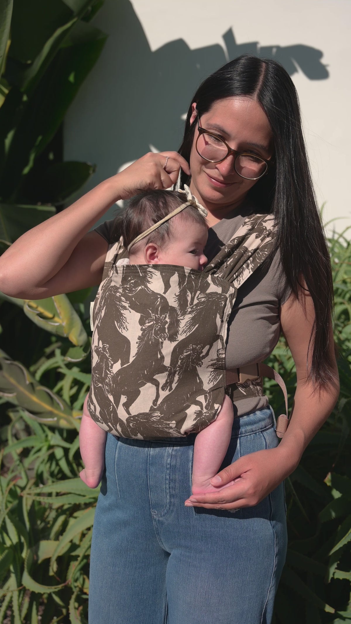 Sprout Newborn Carrier in Rodeo — caregiver in glasses looking down at a newborn in a bow headband, mustang print carrier, aloe plants behind.