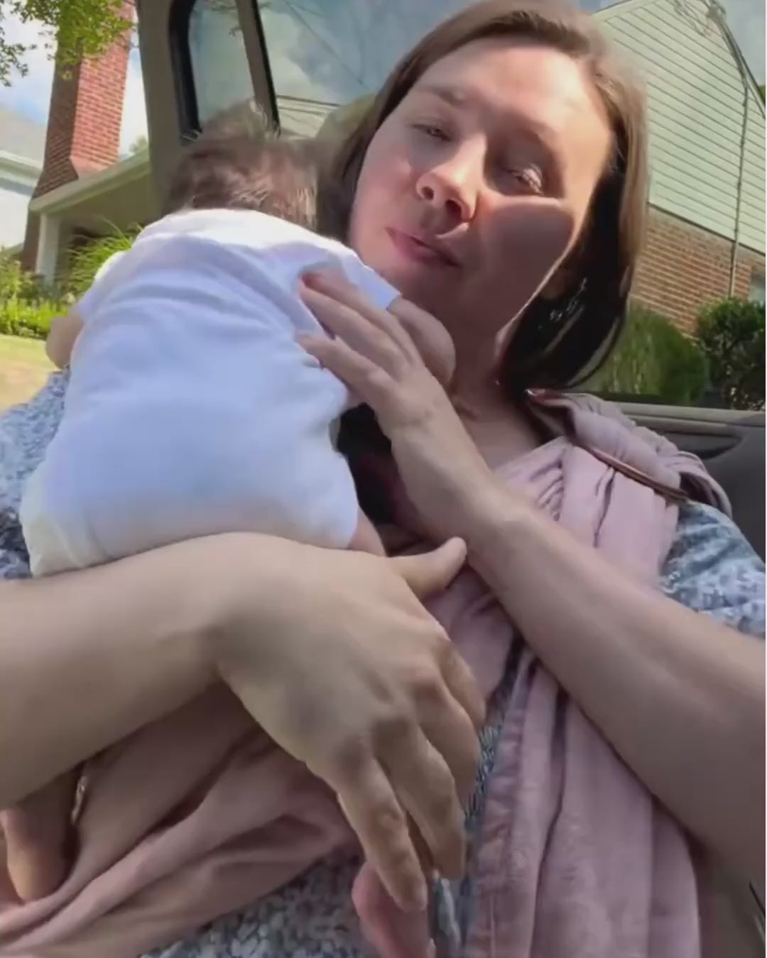 Skye, co-founder of Hope & Plum, settles a newborn baby into a peony ring sling against her chest, adjusting the fabric as she stands outdoors beside a car on a sunny day.