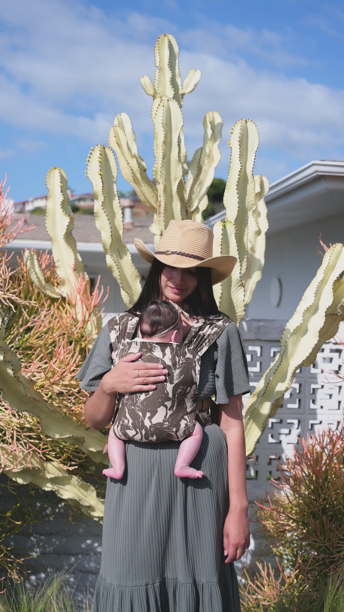 Sprout Newborn Carrier in Rodeo — caregiver in a straw hat and sage dress holding a newborn in the mustang print carrier, cactus behind.