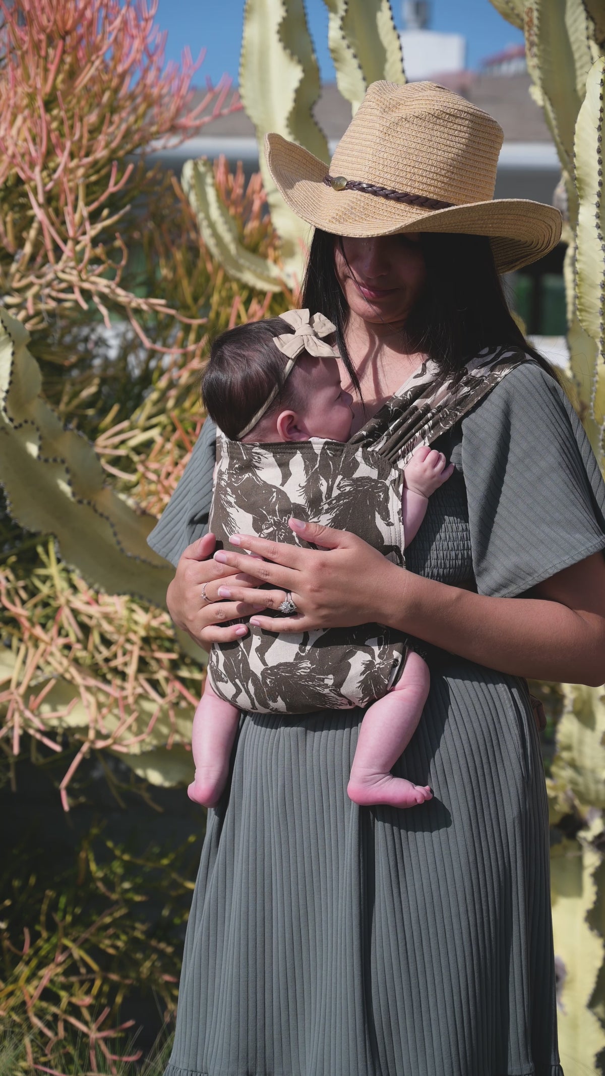 Sprout Newborn Carrier in Rodeo — caregiver in a straw hat, side profile, holding a newborn in a bow headband in the mustang print carrier.
