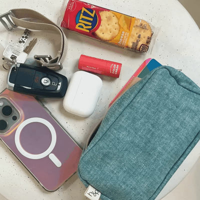 Flat-lay of a Azure Hope & Plum Carrier Pocket pouch alongside everyday essentials it holds: car keys on a strap keychain, a pack of Ritz cheese crackers, a tinted color-cream stick, AirPods, and an iridescent phone, arranged on a cream speckled surface.