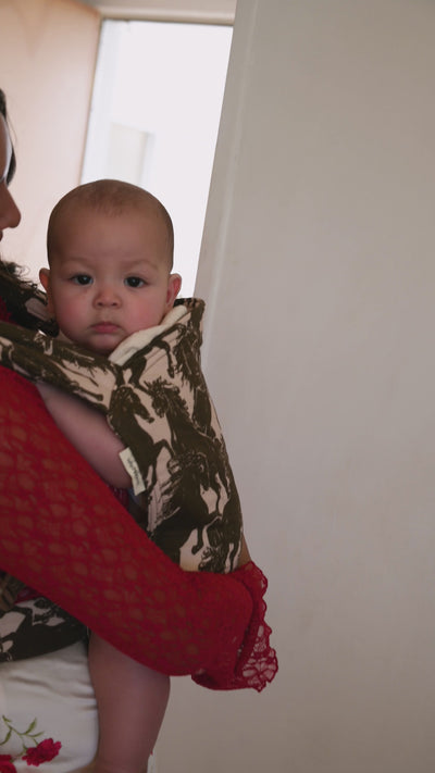Lark Baby Carrier in Rodeo — caregiver in a red lace top embracing an infant in the mustang print carrier, indoors by a window.