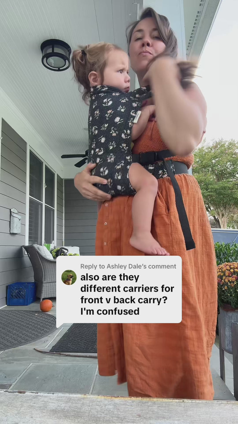 A woman in an orange skirt stands on a covered porch demonstrating how to carry a toddler in a floral-print Lark carrier, mid-gesture as she explains the move. On-screen text reads: 'which I'm gonna show you here' above a reply to a comment asking 'also are they different carriers for front v back carry? I'm confused.'