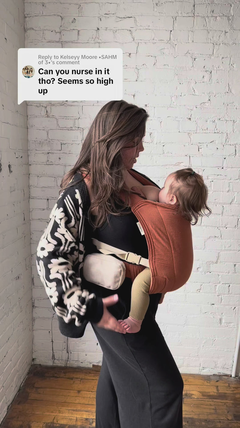 A smiling woman holds her toddler close in a floral-print Lark carrier, the child's head resting against her chest while nursing. On-screen text reads: 'I needed something that I could easily whip a boob out.'