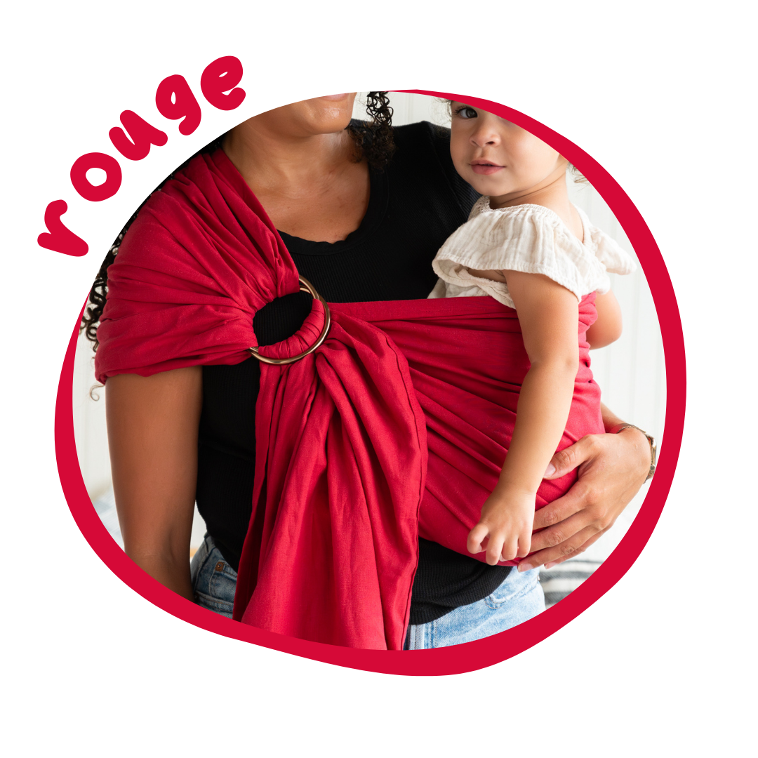 A woman with curly black hair holds her toddler in a hip carry using a ruby red ring sling.