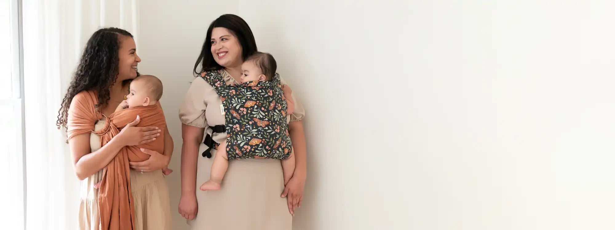Two mothers, one babywearing her baby in a brown ring sling and the other wearing her infant in a baby carrier looking at one another and smiling. 