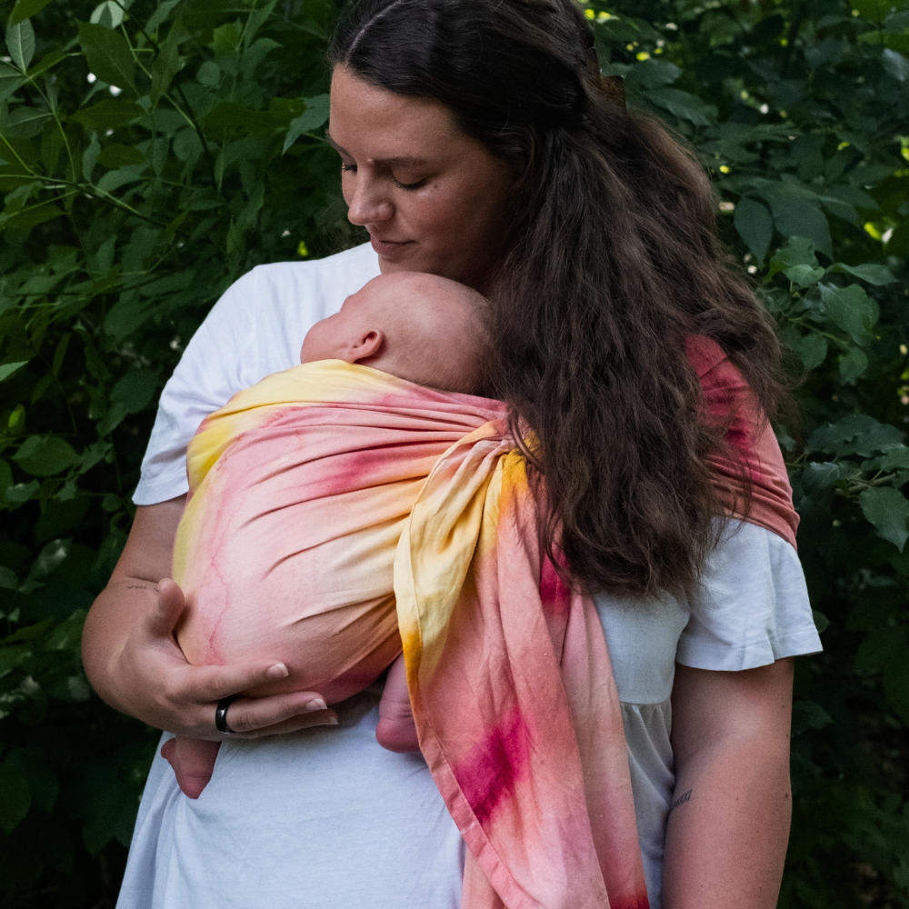 brunette woman looking down at her infant  being carried in a pink and yellow watercolor sling with foliage in the background